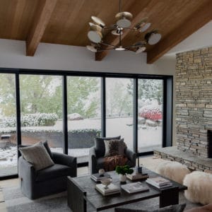 Mid-century modern living room with wood ceiling and beams, a modern large light fixture hanging in the center. Large black framed window wall at the back and a stacked stone fireplace to the right. Leather sofa with pillows in the foreground, a modern dark wood coffee table with decor in the center, and two gray swivel chairs in front of the window.