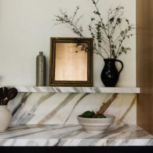 close-up of the bold veined marble countertops above black cabinets. The marble wraps up about 12" to a floating shelf with decor on the floating shelf. A burnished brass picture sconce is on the wall above.