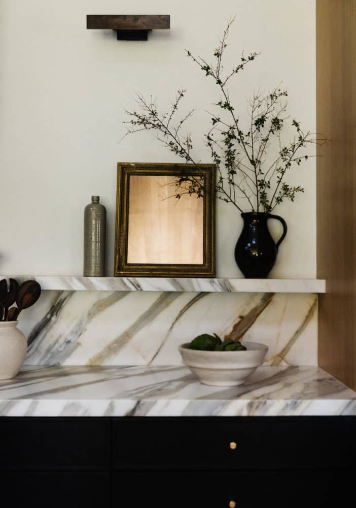 close-up of the bold veined marble countertops above black cabinets. The marble wraps up about 12" to a floating shelf with decor on the floating shelf. A burnished brass picture sconce is on the wall above.