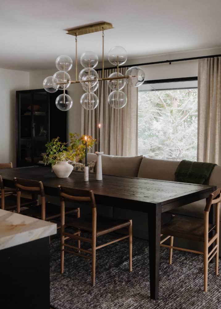 dark wood dining room table with lighter wood chairs on one side with a linen sofa as a bench for the other side. glass and brass bubble light fixture above the table and linen drapes around the large window at the back.