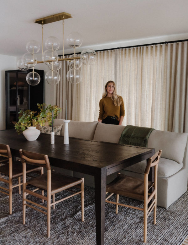 dark wood dining room table with lighter wood chairs on one side with a linen sofa as a bench for the other side. glass and brass bubble light fixture above the table and a woman standing behind the sofa in front of closed linen drapes.