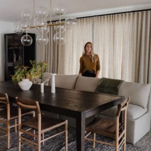 dark wood dining room table with lighter wood chairs on one side with a linen sofa as a bench for the other side. glass and brass bubble light fixture above the table and a woman standing behind the sofa in front of closed linen drapes.