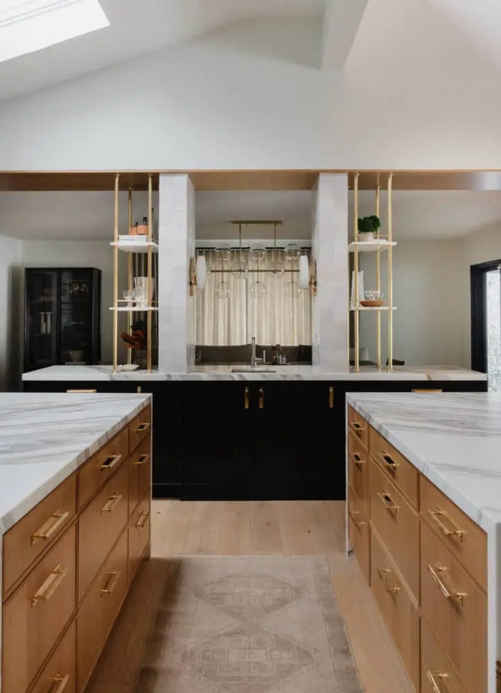 double kitchen islands on each side of the image with white oak cabinets and marble countertops. A black cabinetry bar in the background that acts like a room divider with brass and marble shelves and the dining room in the background