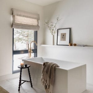 modern squared off freestanding tub with a brass floor mounted faucet with a floating shelf above and a large window to the left with a linen roman shade.