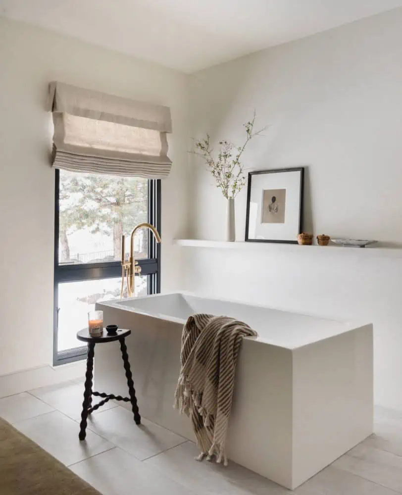 modern squared off freestanding tub with a brass floor mounted faucet with a floating shelf above and a large window to the left with a linen roman shade.