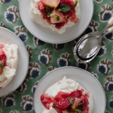 mini strawberry pavlovas on small ceramic dishes on a green and blue floral placemat with spoons.