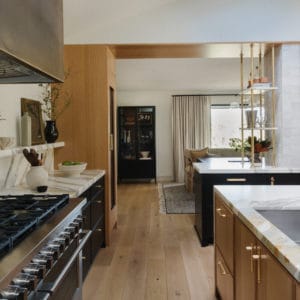 kitchen image with the oven wall to the left and an island to the right. The island has white oak wood cabinets and the oven wall has black cabinets with marble countertops that wrap up to a floating shelf. The dining room is in the background.