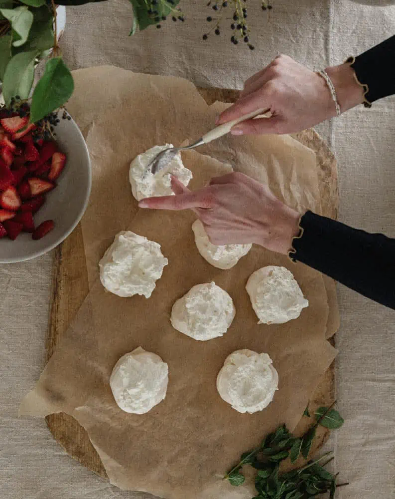 top view of womans hands adding whipped cream to the top of mini pavlovas sitting on top of parchment sheets with a bowl of strawberries in the corner and mint leaves on the table.