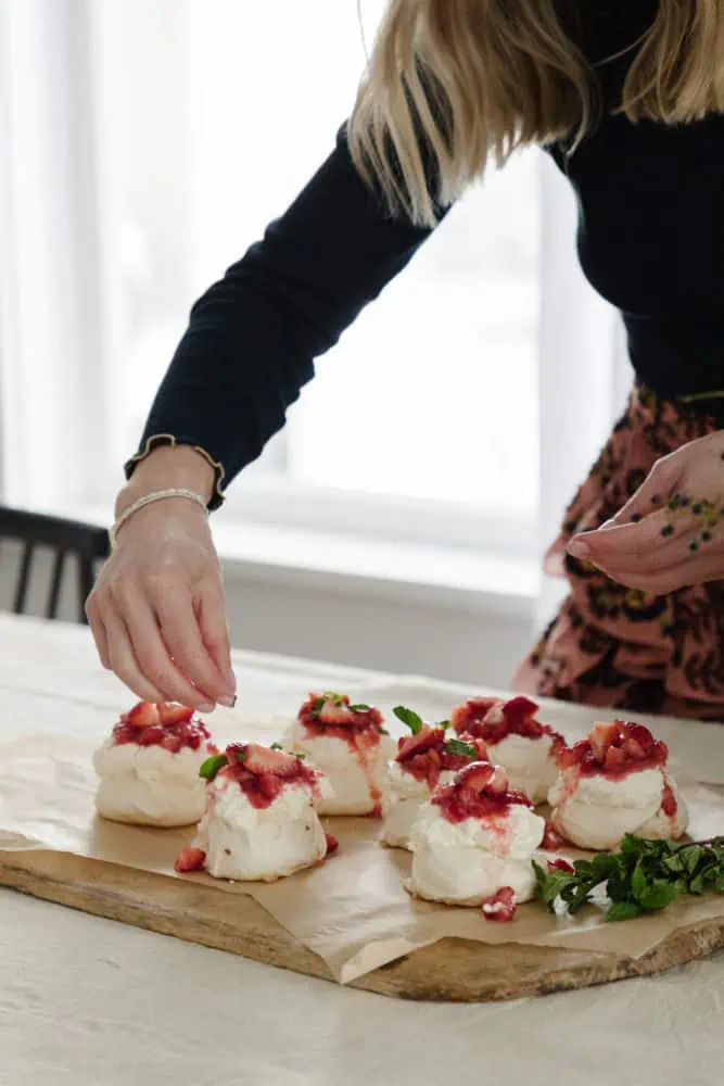 woman's hand adding mint leaves to the top of mini pavlovas topped with whipped cream and strawberries with a large wooden cutting board below.