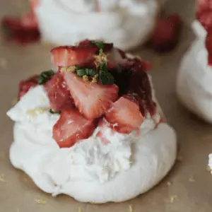 close up photo of a mini pavlova topped with whipped cream, strawberries, mint, and lemon zest.