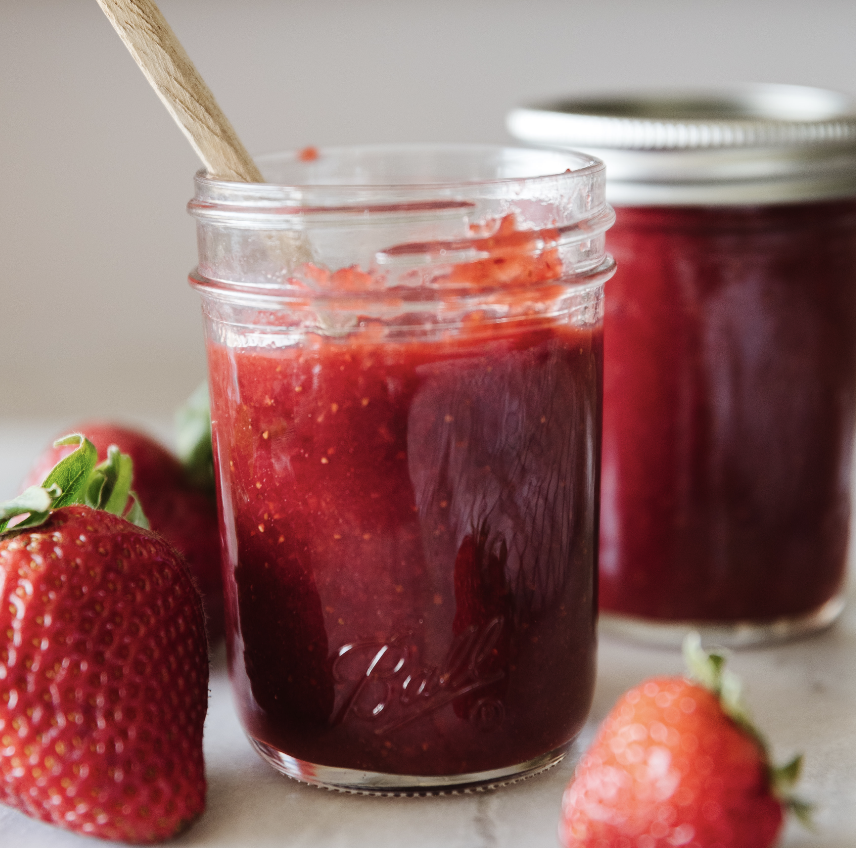 glass mason jar filled with strawberry honey butter with a wooden spoon sticking out of it. Fresh strawberries in the foreground and another closed jar of strawberry honey butter in the background.