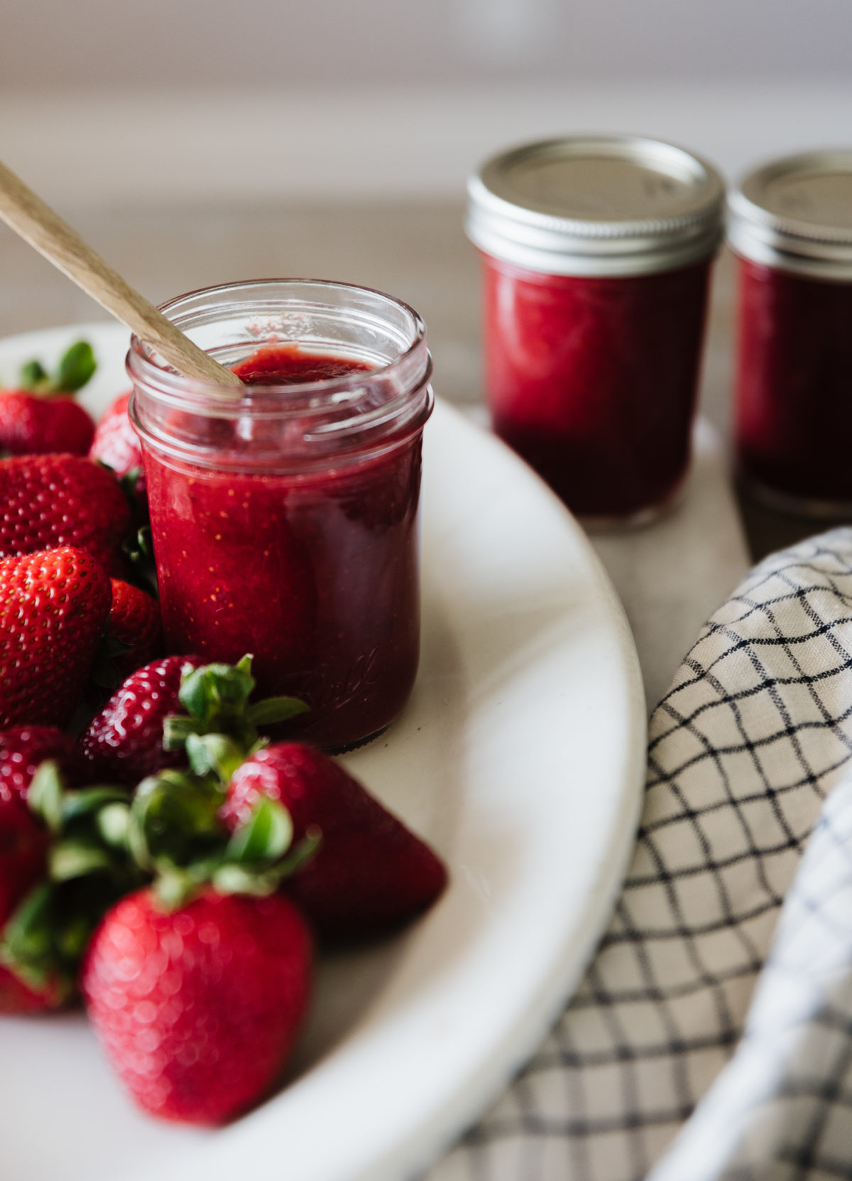 glass mason jar filled with strawberry honey butter with a wooden spoon sticking out of it on a white oval platter surrounded by fresh strawberries in the foreground and two closed jars of strawberry honey butter in the background.