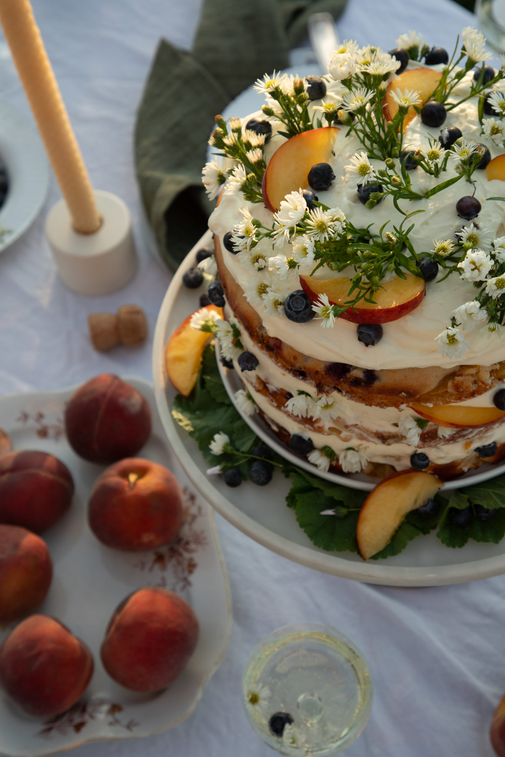 a close up photo of a a cake with whipped cream cheese frosting topped with peach slices, blueberries, and camomile flowers with peaches on the table in front of the cake