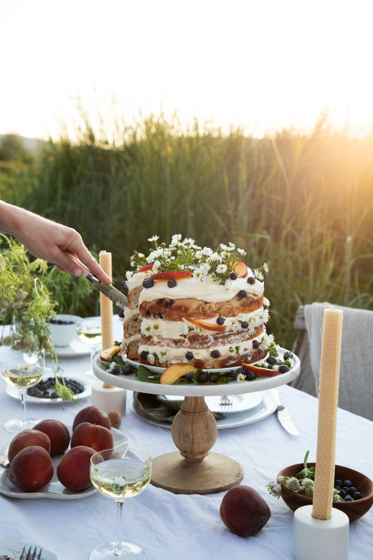 a woman's hand cutting into a cake that's topped with peaches, blueberries, and camomile flowers on a white cloth covered table in a field with peaches and candlesticks on the table as well.
