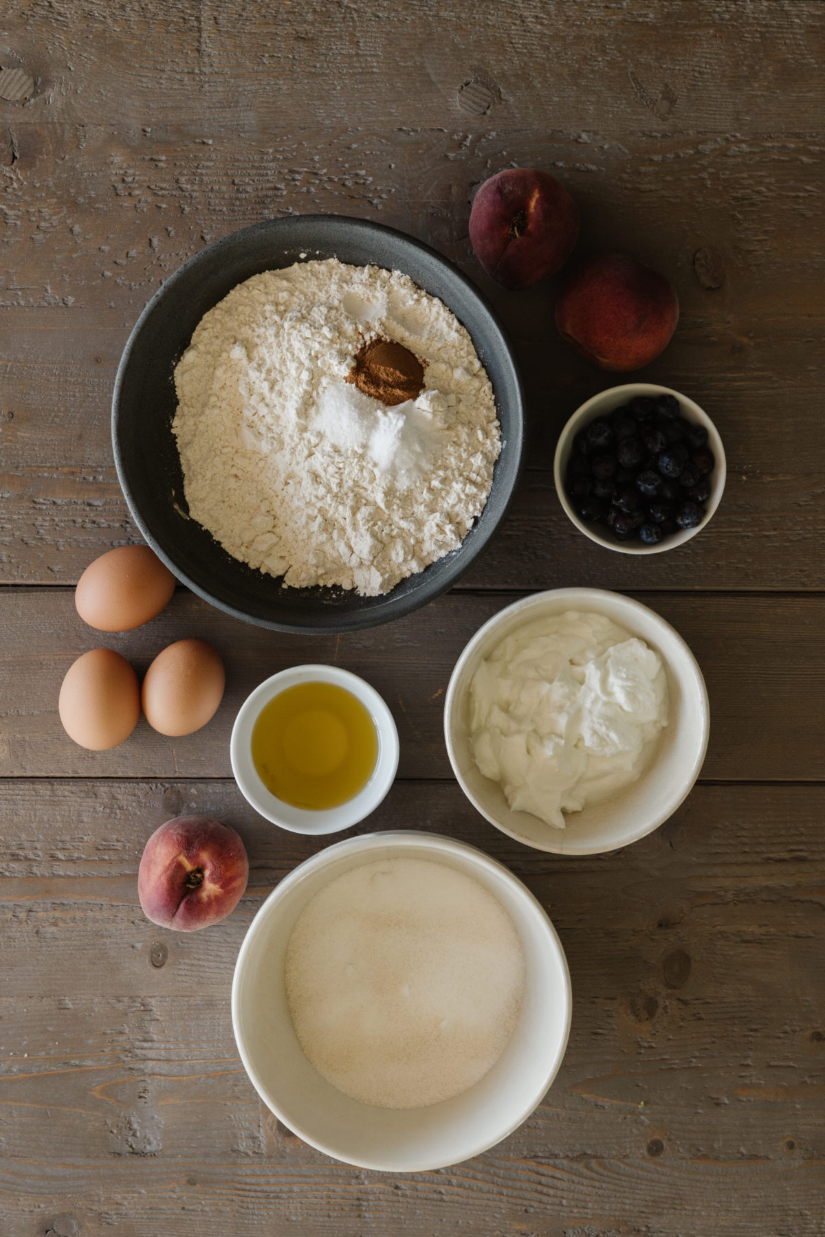 peach cake ingredients in various neutral colored bowls, 3 brown eggs, and some peaches on a rustic wooden table top