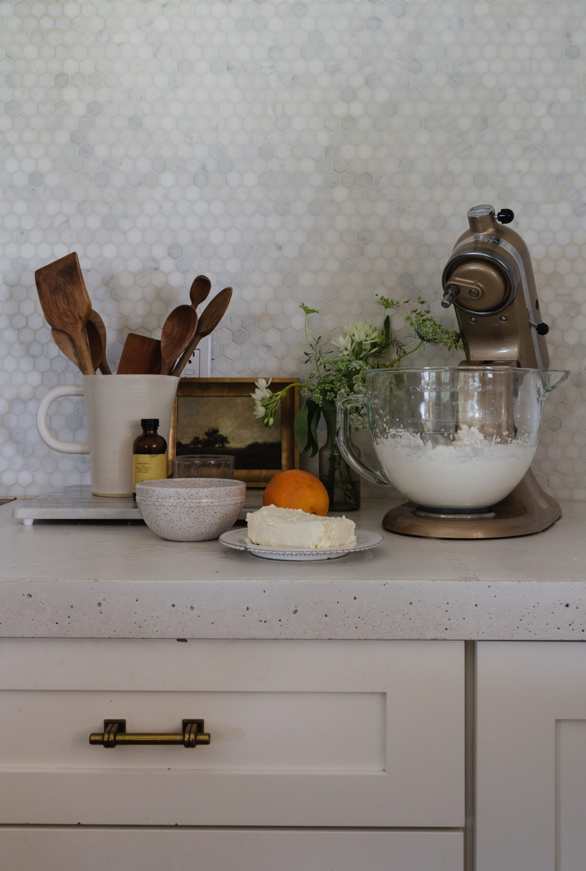 white kitchen with marble penny round backsplash and white concrete countertop with a white jar filled with wooden utensils, artwork, flowers, and the ingredients for cream cheese frosting next to a gold kitchen aid mixer.