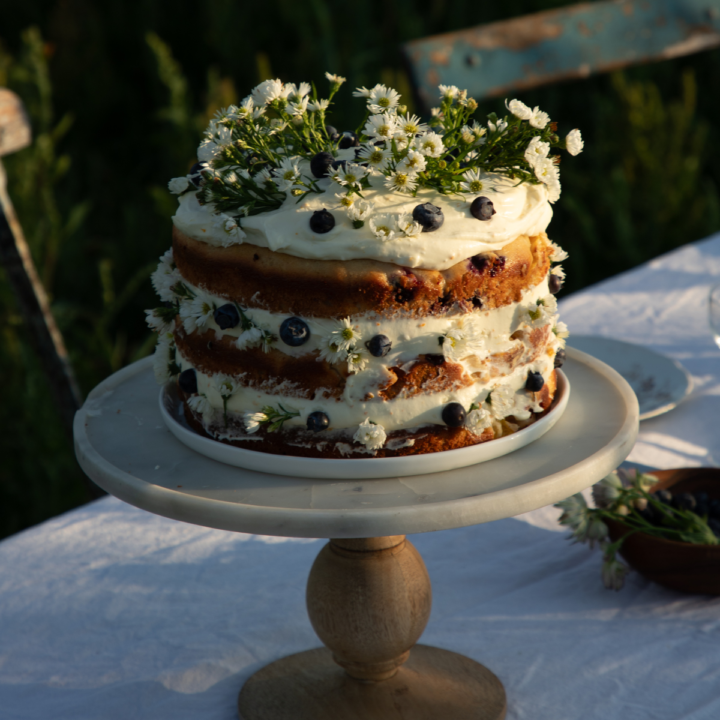 peach cake with orange zest whipped cream cheese frosting on a marble and wood cake stand on a white cloth covered table in an outdoor setting.