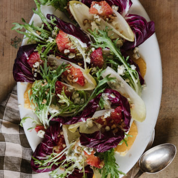 A vibrant endive salad presented on a white plate, featuring crisp Belgian endive and radicchio leaves arranged in a circular pattern. The salad is topped with bright orange slices, crumbled blue cheese, and crunchy walnuts. Fresh parsley adds a touch of green, creating a beautiful and appetizing dish perfect for any gathering.